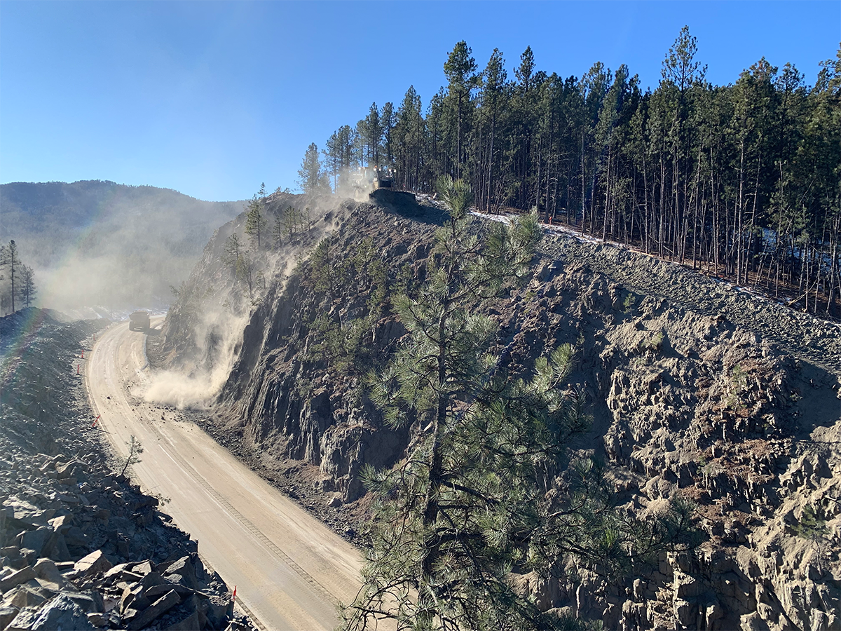 Machines doing earth work on top of a steep slope. Debris (rocks, dirt) falling down the slop onto the roadway below.