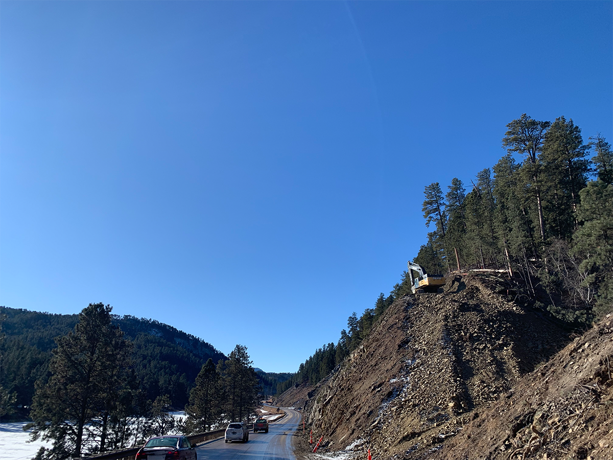Cars being led by a pilot car while machine does earthwork on the adjacent slope.